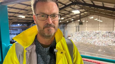 Councillor Chris Watts, cabinet member for the environment and transport stands in front of a mountain of waste at a recycling plant. 