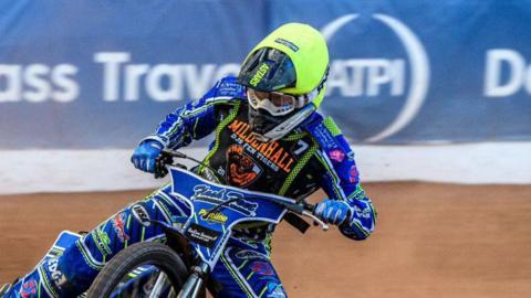 A speedway racer steers his bike around a corner on a mud track. He wears a bright green helmet and blue racing suit with various sponsors written on it.