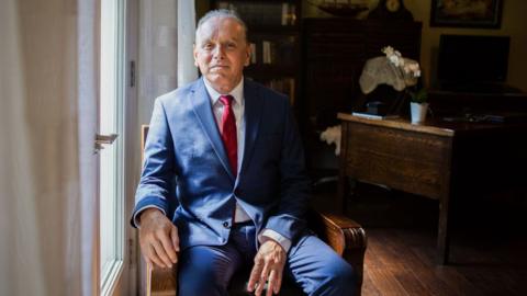 Dr Marrero, in a navy suit and red tie, sits in a wooden chair in his home office, in the light coming in from the door windows