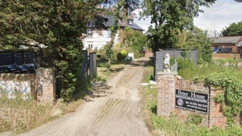 Exterior of a large white house surrounded by a brick wall on either side of the house. There are bushes and small statues at the front of the house with a large tree hanging over the front. There is a small black sign at the front with white writing saying Peter House and the telephone number.