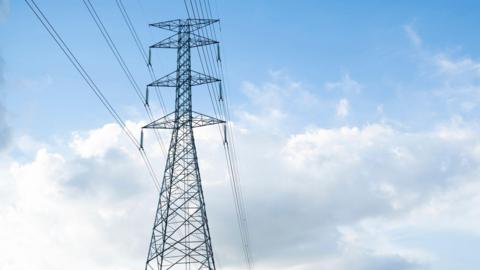 A large electrical tower with power lines coming from both sides of it. The background is a blue sky with white clouds. 