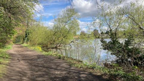 A view of Poynton Pool on a sunny spring day. The image shows the pool on the right, with a footpath on the left. There are a number of trees along the bank of the pool.