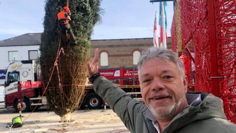 Man pretending to hold up a wonky Christmas tree with his hand.