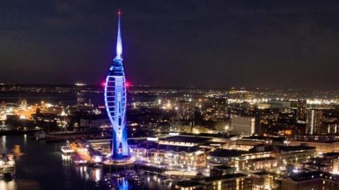 View across Portsmouth city at night. Buildings are lit up with lights whilst the sky is dark. In the foreground is the Spinnaker Tower, an iconic tall structure in the shape of a ship's sale which is illuminated blue.