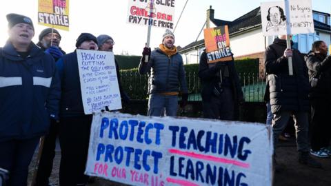 A group of people in winter clothing hold up sign supporting the strike action. A sign in the foreground reads: "Protect teaching, protect learning."