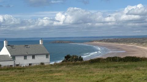 White clouds across a blue sky with sea, sand and a house below 