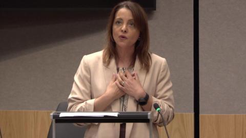 Shav Taj holding her hands to her chest, speaking and looking upwards. She is wearing a beige jacket and is in front of a lecturn.