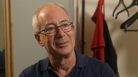 The comedian Ben Elton, a man with short grey hair and glasses, wearing a dark blue polo shirt, looks at something behind the camera.