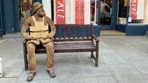 A wicker figure depicting a bearded RNLI crewman from 1886 - including a helmet and lifejacket - is sitting on a bench outside a shop in a town centre. The figure has its hands resting on its legs and is looking to its left.