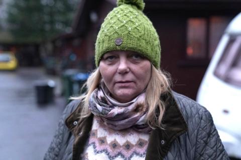 A woman in her early 50s with blonde hair and a green woolly hat. She is wearing a black padded jacket, scarf, and a patterned jumper.
