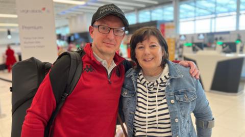 Steve wears a red jumper, black hat and carries a black rucksack. He has his arm around sue who wears a denim jacket over a blue and white striped jumper. She has bobbed dark hair.