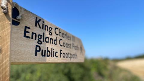 A wooden sign which reads "King Charles III England Coast Path" stands on a grassed area with sand and blue sky visible in the background.