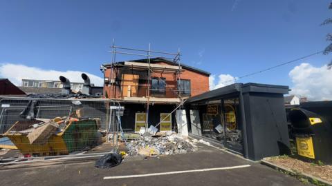 A restaurant destroyed by an arson attack. A small two storey-building is seen with rubble piled in front of it, black staining on the red bricks from the smoke and scaffolding up against the building. A skip is also outside the premises piled with rubbish.