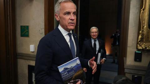 An image of Mark Carney in the foyer of Canada's House of Commons. He is seen speaking to a person off-camera while holding a document titled 'Un Canada Fort Pour Tous'. Beside him is finance minister Francois-Philippe Champagne, who is looking over at Carney and is holding the same document.