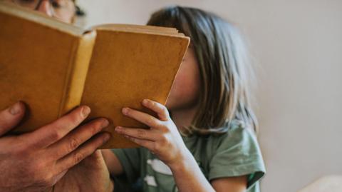 A child reading a book with an older person. The book is covering the faces of both people. 