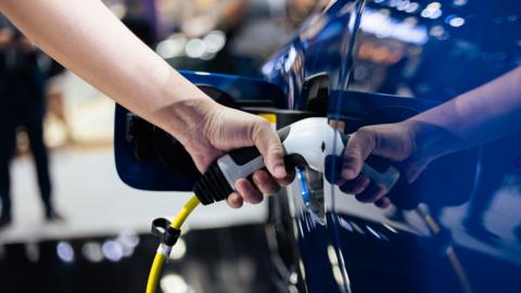 An arm plugging in yellow-wired white-headed charging cable into a blue electric car 