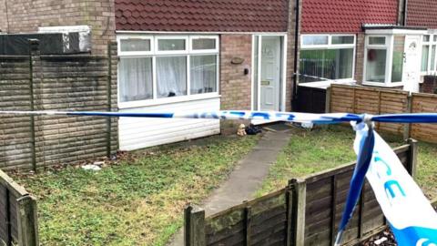 A blue police cordon blocks the entry to a pale-bricked house. The tape stretches across the image which shows a property with a narrow concrete path leading to a white plastic door that is partially open. There is grass on either side of the path and a low fence at the front of the house.