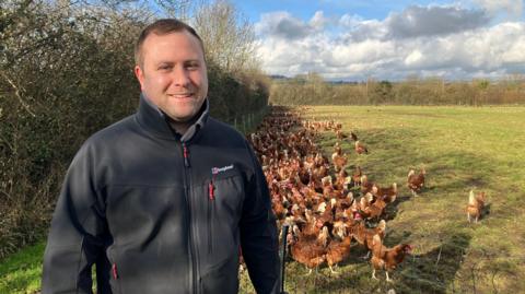 Tom Wood shown wearing a grey Berghaus fleece in a field, smiling at the camera. There are hundreds of hens behind him, gathered next to a hedge. 