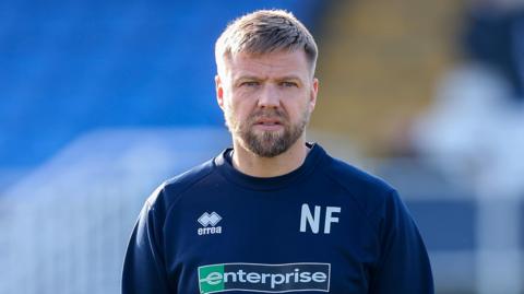 Nicky Featherstone in a navy blue tracksuit with 'NF' in the left chest area, stood with hands in pockets ahead of a Hartlepool home game 