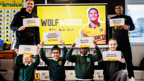 Joao Gomes is dressed in a black and gold Wolves tracksuit, stands behind a row of boys and girls with another male club representative. They are all holding up motivational sentences about speech. There is a poster of Gomes and the words "Wolf Words" behind them all. The pupils are in green and green school uniform. 