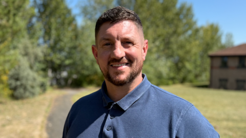 Colin Burns, who has brown gelled hair and a brown beared, is wearing a dark blue button-down T-shirt. He is smiling in front the camera as he stands on a concrete path. In the background, there are tall trees and faded grass. To his right, there is an office building which has a red roof and beige coloured bricks.