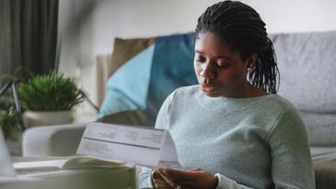 A woman sat on a living room floor looking at a heating bill. She is wearing a light green jumper and behind her is a grey sofa, a plant on a table, and a white wall.