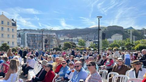 A picture of people gathering for Liberation Day in 2024. There is people sat in a crowd wearing hats and there are Jersey flags around the crowd. There is white chairs and buildings in the distance.