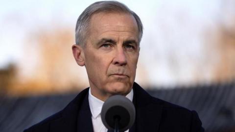 Canadian Prime Minister Mark Carney speaks during a press conference in Beijing, China, 16 January 2026. He is seen standing in front of a microphone, and is wearing a black coat over a suit and tie. He has a serious expression on his face. He has short black and grey hair that is parted to the left. 