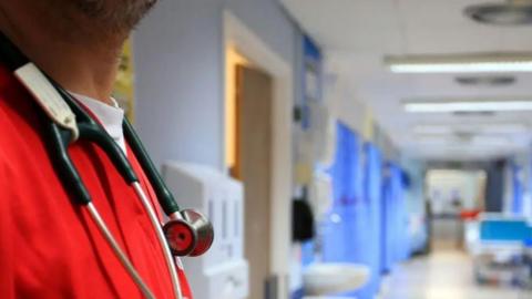 A man in red scrubs with a stethascope stands at the end of a hospital ward you can only see his chest. Blurred in the background you can make out a corridor with blue curtains, a sink and a hospital bed.