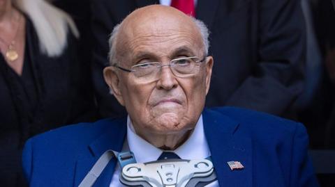 Giuliani wearing a brace on his chest. He has a blue suit and glasses and looks towards the camera