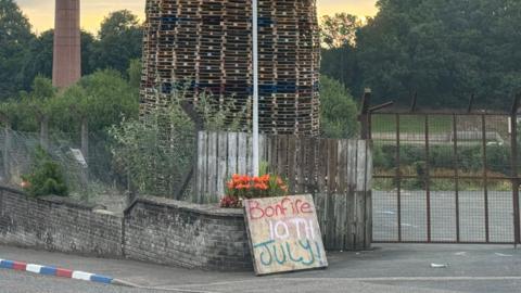 A bonfire of wooden pallets sits behind a fence, in front of the fence is a brick wall and a wooden sign is propped up against it with "bonfire 10th July" written on it