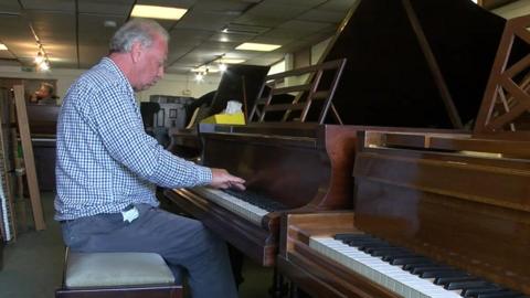 David has a blue-checked collared shirt and blue trousers. He has short grey hair. He is playing a wooden grand piano in the middle of a row of pianos.