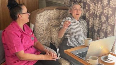 A member of care home staff in a pink tabard sits next to an elderly lady who is sat at a table with a laptop and cup of tea as well as a plate of biscuits.