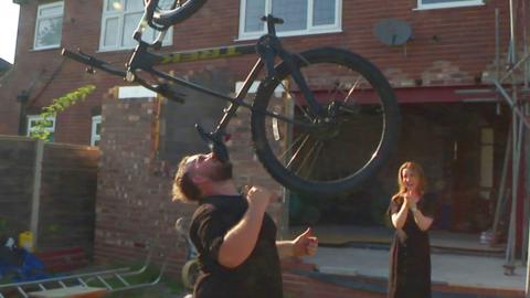 A man balances a bike on his chin outside a house as a woman laughs in the background
© BBC