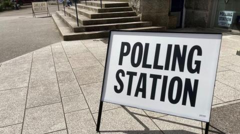 A metal sign on a stand saying "Polling Station" on a small plaza next to some grey stone steps.