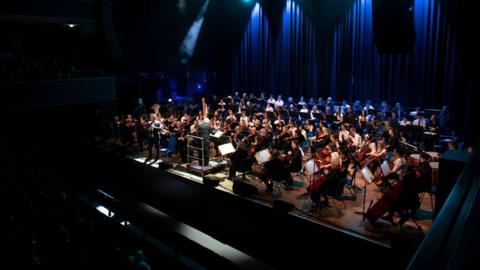 A large orchestra performing on a stage in front of an audience. Blue curtains can be seen behind the stage. The stage is bathed in light while audience members sit in the dark.