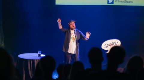 A man wearing a baseball cap and glasses stands on stage in front of a crowd of people, performing spoken word poetry. The backdrop is blue.