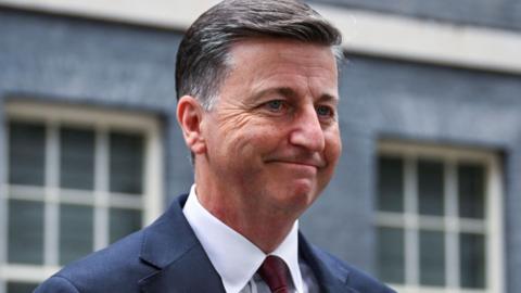 Douglas Alexander wearing a dark blue suit, white shirt and dark red tie walks out of 10 Downing Street.