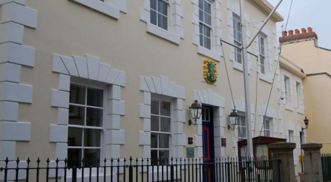 Alderney's Court building exterior - A yellow building with an iron fence in front of it. In front of it is also a pole for a sail. 
