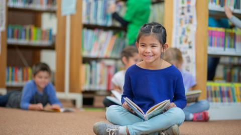 A young girl wearing a blue top and blue trousers sits cross legged on the floor. She has an open book on her lap and is smiling. In the background a young boy and two girls are also sat with open books on the ground. There are shelves of books behind them.