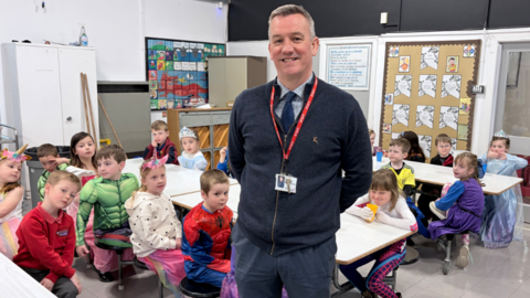 In a school canteen, a man in a navy zipped cardigan and tie with a red lanyard is standing with his hands behind his back in front of some primary school children in fancy dress sitting at white tables