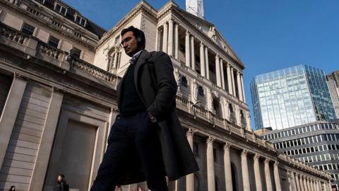 A man in a long dark coat is striding past the Bank of England building on a sunny day.