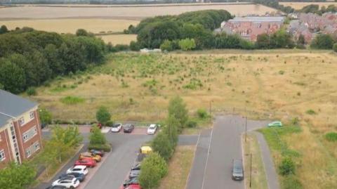 Drone shot of a patch of in between housing estates, with fields off into the distance. Cars parked outside one building.