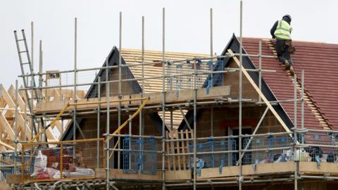 Houses being constructed. The top part of two roofs is visible. The wooden beans are showing and a person is standing on one roof. 