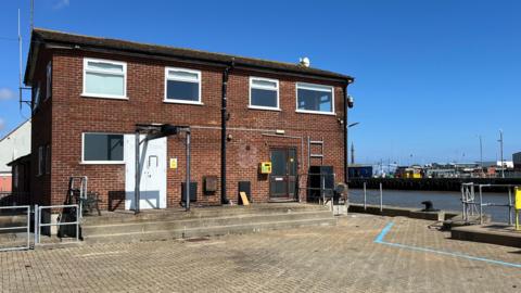 The Great Yarmouth River Port Pilots' Station in Gorleston is an red brick building of the 1970s, with a number of white-framed windows, and a shallow-pitched roof. In the foreground is brick-weave paving, with markings on the ground, and some hand rails and steps down to the river. Beyond the river, on the opposite back, are a series of port buildings and structures.