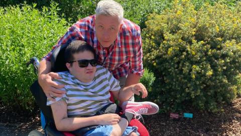 A man with white hair stands next to a younger man sitting in a wheelchair wearing sunglasses.