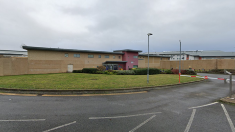 The outside of HMP Peterborough. A pale-bricked building with a part rendered and painted red. There is a green piece of land in the front.  