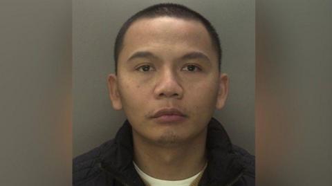A mug shot of Roman Le. He has short dark hair and wears a dark coat and a white T-shirt.
