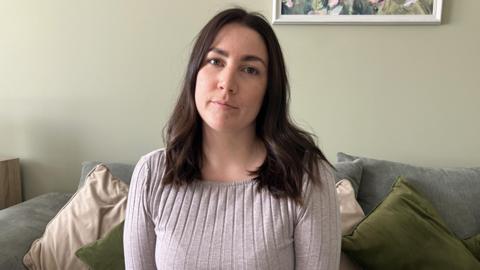 A woman with dark, shoulder-length hair sat on a sofa in her living room wearing a grey long sleeved T-shirt. 