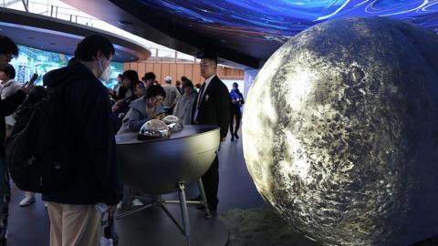 Visitors look at moon samples encased on a table at an expo. There is a large sculpture of the moon near them.